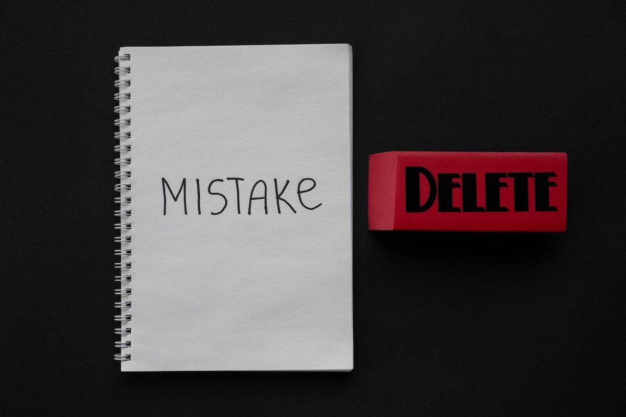 Notebook with 'MISTAKE' written on open page and adjacent eraser labelled 'DELETE' on black tabletop