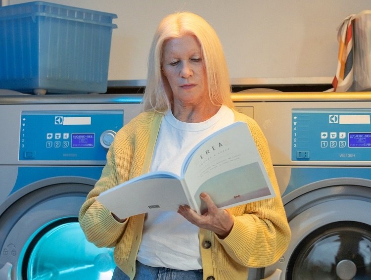 Blonde woman wearing white and yellow leaning against washer and dryer white reading