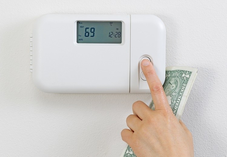 Hand holding dollar bill while adjusting temperature settings on thermostat
