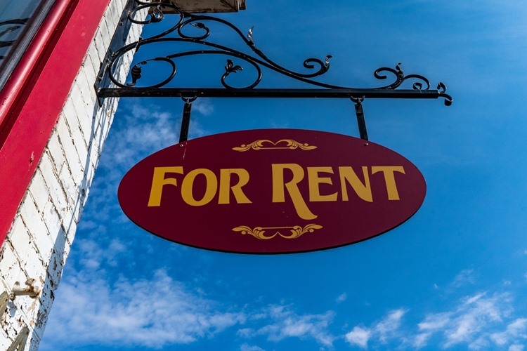 Hanging 'FOR RENT' sign on white brick building with red window trim