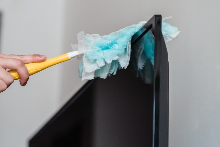 Blue and yellow duster removing dust from upper edge of flat-screen television in white living room